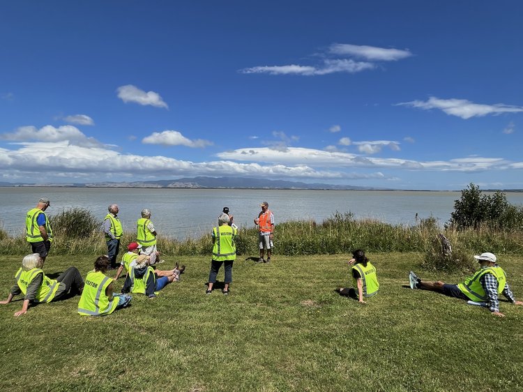 A group of atendees in high-vis vest sitting by the edge of the Wairarapa Moana listening the Dr Nicola Litchfield talk about the geological history of the lake.